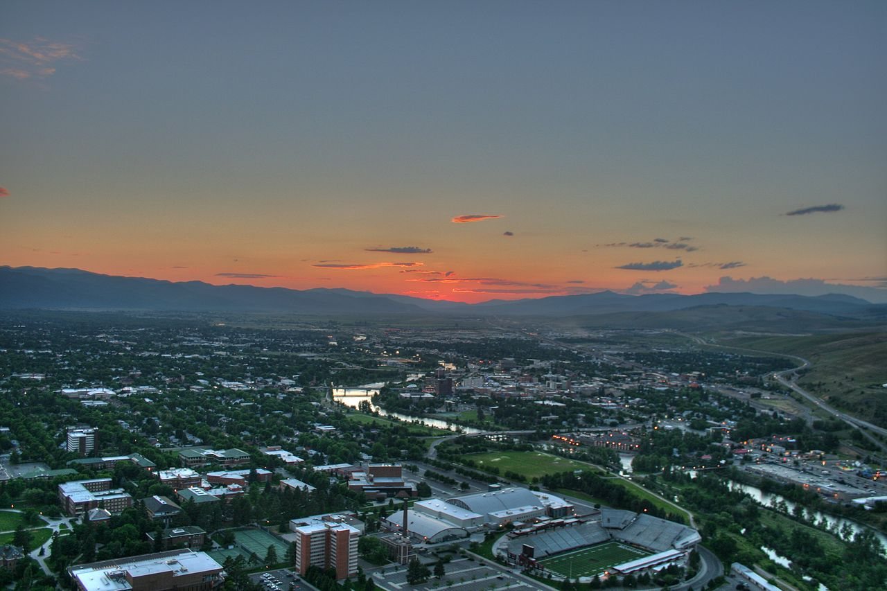 Missoula at dusk from above, with the Clark Fork River running through town and mountains on the horizon
